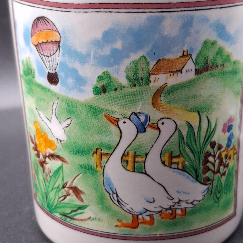May include: White ceramic mug with a pastoral scene. The design features two geese wearing hats, a hot air balloon, a house, and a winding path. The mug has a pink border and is decorated with colorful illustrations.