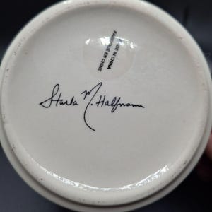 K&ouml;nnte beinhalten: Der Boden einer wei&szlig;en Keramiktasse mit der Unterschrift "Starla M. Halfmann" in schwarzer Schrift. Ein klarer Aufkleber mit dem Text "MADE IN CHINA" und "FABRIQUE EN CHINE" ist sichtbar.