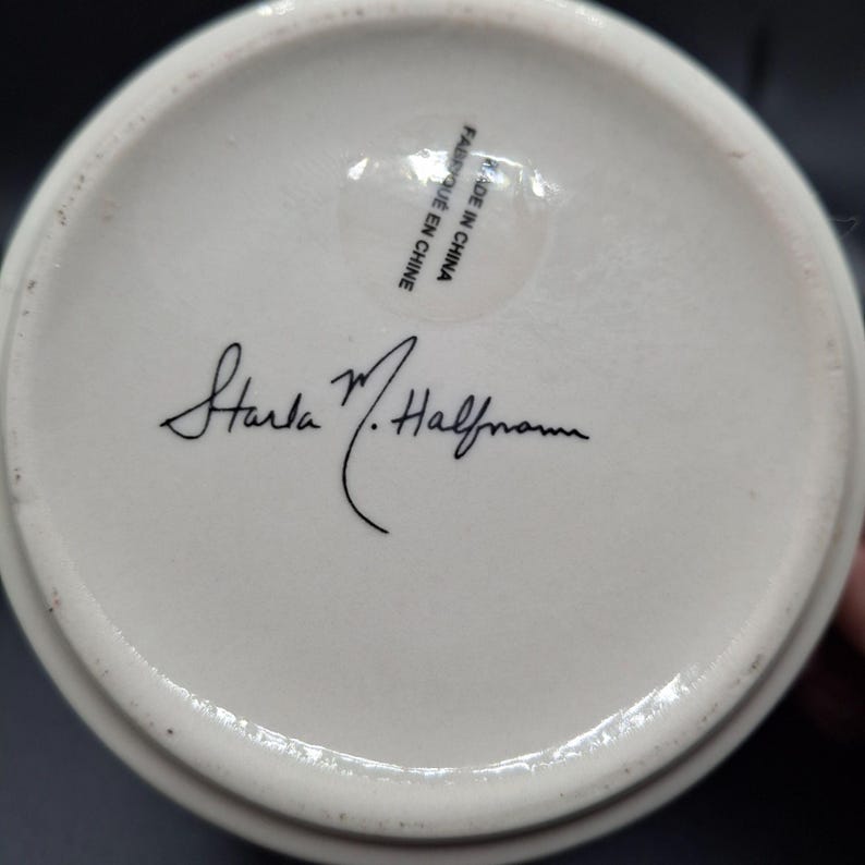 K&ouml;nnte beinhalten: Der Boden einer wei&szlig;en Keramiktasse mit der Unterschrift "Starla M. Halfmann" in schwarzer Schrift. Ein klarer Aufkleber mit dem Text "MADE IN CHINA" und "FABRIQUE EN CHINE" ist sichtbar.