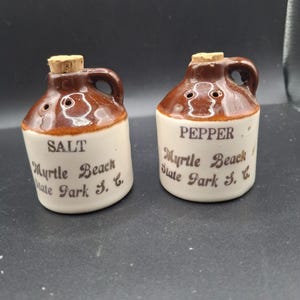 Op de afbeelding: Een paar keramische zout- en peperstrooiers in de vorm van miniatuurkruiken. De strooiers zijn bruin en wit met kurken stoppers en handvatten. De tekst "SALT" en "PEPPER" staat op de voorkant, samen met "Myrtle Beach State Park S.C."