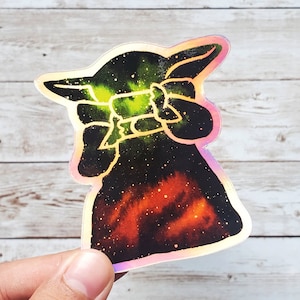 May include: Holographic sticker featuring a stylized character with a space-themed design. The sticker has a black background with green and orange nebula effects and a light outline. The character is holding a small object.