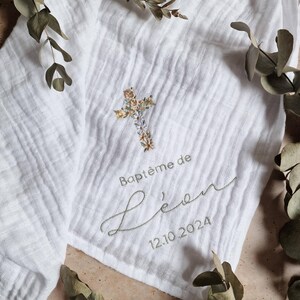 Op de afbeelding: Witte mousseline doek met geborduurd bloemenkruis en tekst. De tekst luidt "Baptême de Léo 12.10.2024" in groene draad. Eucalyptustakken zijn rond de doek geplaatst.