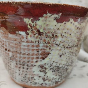 May include: A handmade ceramic bowl with a textured exterior and a rich color palette. The rim is a deep red, transitioning to a lighter, speckled pattern of white, brown, and yellow. The bowl's design features a grid-like texture.