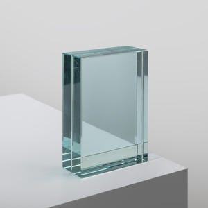 Puede incluir: Un bloque rectangular hecho de vidrio transparente con un tinte azul claro. El objeto presenta bordes gruesos y pulidos con una superficie lisa y reflectante, manteniéndose en posición vertical con una apariencia sólida y líneas nítidas.