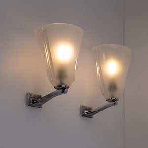 May include: Two wall-mounted sconces with frosted glass shades and chrome-finished arms. Each sconce has a bright light emanating from within the shade. The sconces are mounted on a white wall, creating a clean and modern aesthetic.