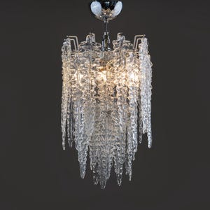 May include: A clear glass chandelier with a chrome finish. The chandelier has a unique design with icicle-like glass elements hanging from a rectangular frame. The light fixture is suspended from a chrome canopy.