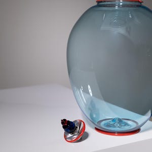 May include: A large, blue glass vase with a red rim and base. A smaller, decorative bottle stopper with red, blue, and silver accents sits nearby. The vase has a bulbous shape and a smaller top section.
