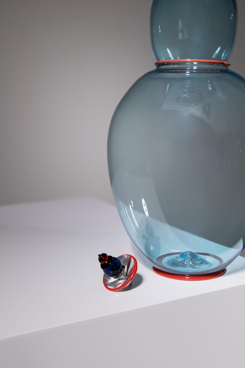 May include: A large, blue glass vase with a red rim and base. A smaller, decorative bottle stopper with red, blue, and silver accents sits nearby. The vase has a bulbous shape and a smaller top section.
