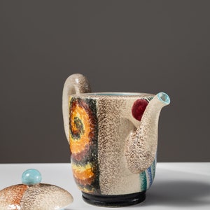 May include: A ceramic teapot with a colorful abstract design in shades of brown, yellow, blue, and red. The teapot has a lid with a blue knob and a spout with a blue rim.