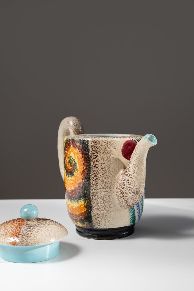 May include: A ceramic teapot with a colorful abstract design in shades of brown, yellow, blue, and red. The teapot has a lid with a blue knob and a spout with a blue rim.