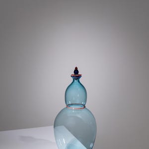 May include: A light blue glass vase with a bulbous lower section and a smaller, rounded top. The vase has a red rim and a red and blue stopper. The vase is displayed on a white surface.