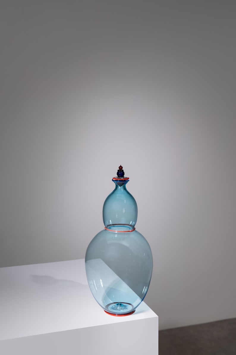 May include: A light blue glass vase with a bulbous lower section and a smaller, rounded top. The vase has a red rim and a red and blue stopper. The vase is displayed on a white surface.