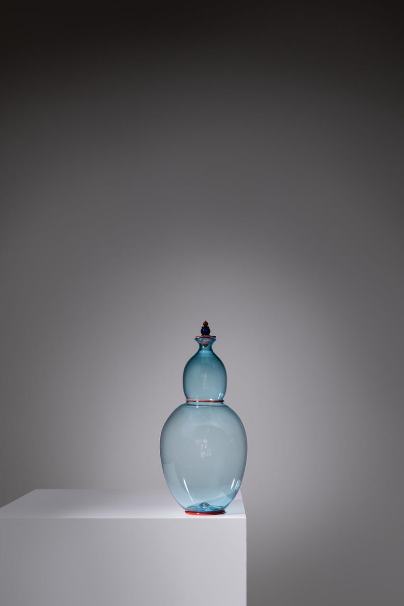 May include: A light blue glass decorative vase with a bulbous lower section and a smaller, rounded upper section. The vase has a red rim and a red and blue stopper. The vase is displayed on a white surface against a gray background.