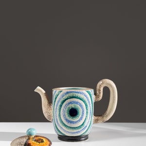 May include: A ceramic teapot with a blue, green, and white eye design. The teapot has a matching lid with a blue, orange, and brown design.