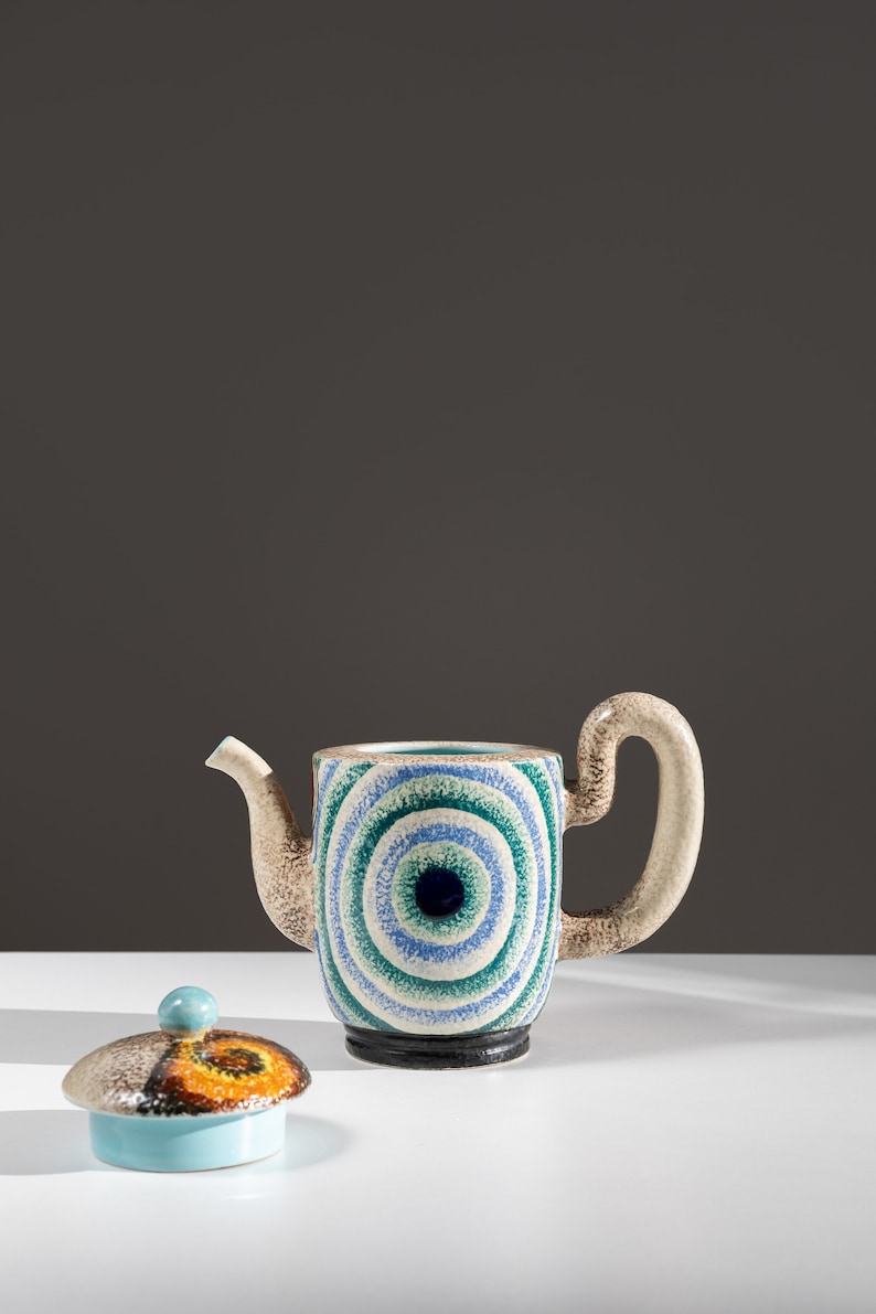 May include: A ceramic teapot with a blue, green, and white eye design. The teapot has a matching lid with a blue, orange, and brown design.