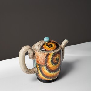 May include: A ceramic teapot with a brown, yellow, and blue swirl pattern. The teapot has a rounded body and a spout on the side. The lid has a small blue knob.