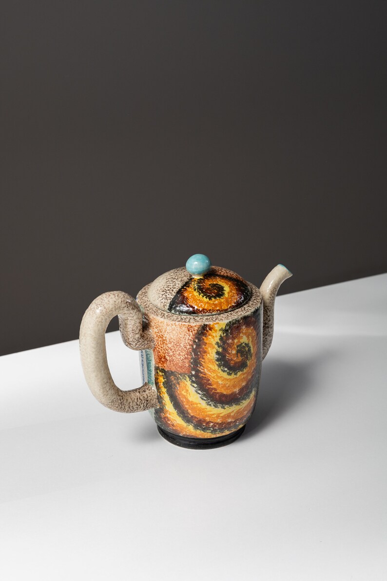 May include: A ceramic teapot with a brown, yellow, and blue swirl pattern. The teapot has a rounded body and a spout on the side. The lid has a small blue knob.