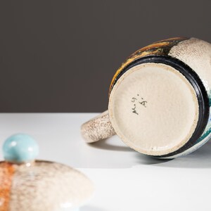 May include: A ceramic teapot with a blue, green, and brown glaze. The teapot has a lid with a blue knob and the bottom of the teapot is marked with the text "M. 59. A."