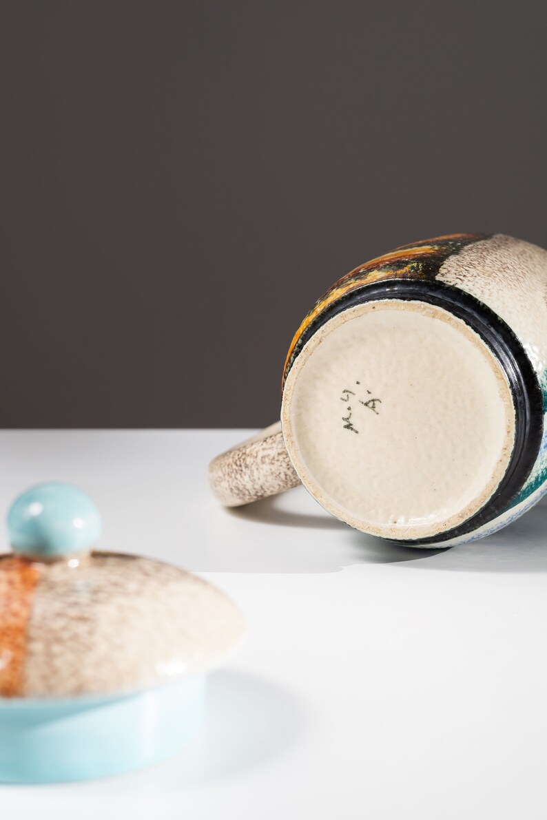 May include: A ceramic teapot with a blue, green, and brown glaze. The teapot has a lid with a blue knob and the bottom of the teapot is marked with the text "M. 59. A."