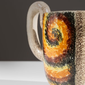 May include: A ceramic teapot with a brown, yellow, and black abstract design. The teapot has a unique handle and a black base.