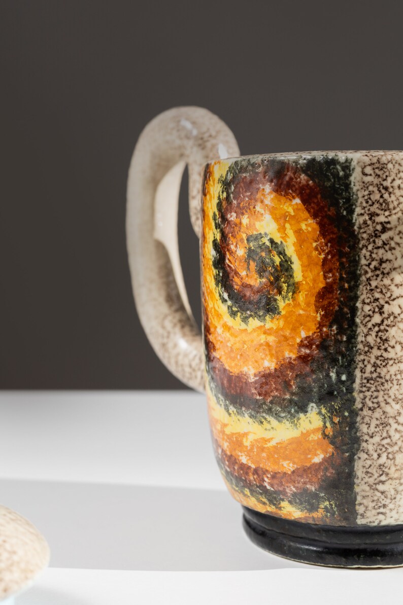 May include: A ceramic teapot with a brown, yellow, and black abstract design. The teapot has a unique handle and a black base.