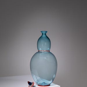 May include: A large, light blue glass vase with a bulbous base and a smaller, rounded top. The vase has a red rim at the base and top. A small, decorative stopper with red and blue accents sits nearby. The vase is approximately 12 inches tall.