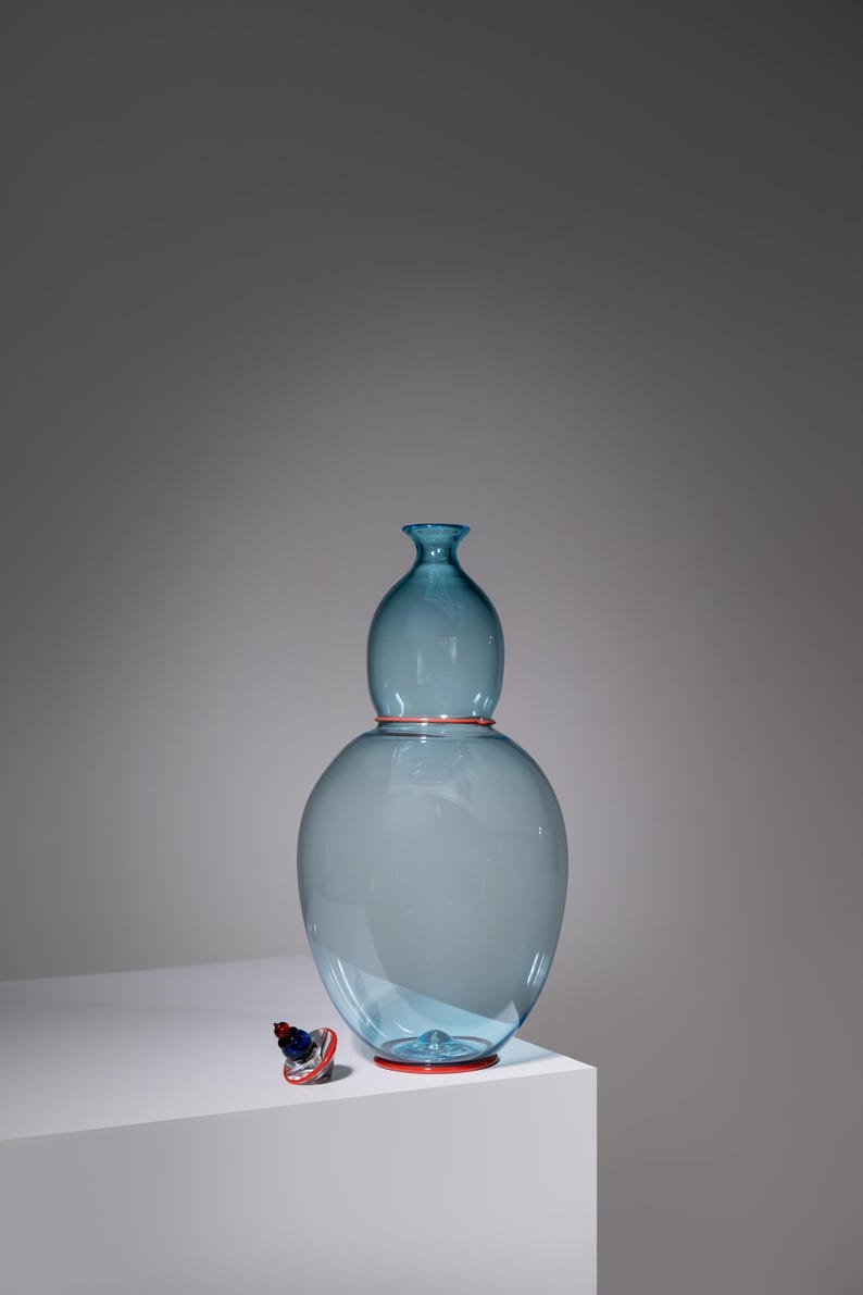 May include: A large, light blue glass vase with a bulbous base and a smaller, rounded top. The vase has a red rim at the base and top. A small, decorative stopper with red and blue accents sits nearby. The vase is approximately 12 inches tall.