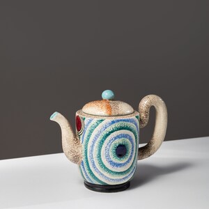 May include: A ceramic teapot with a blue, green, and white geometric pattern. The teapot has a rounded body and a curved spout. The lid has a small, round knob.