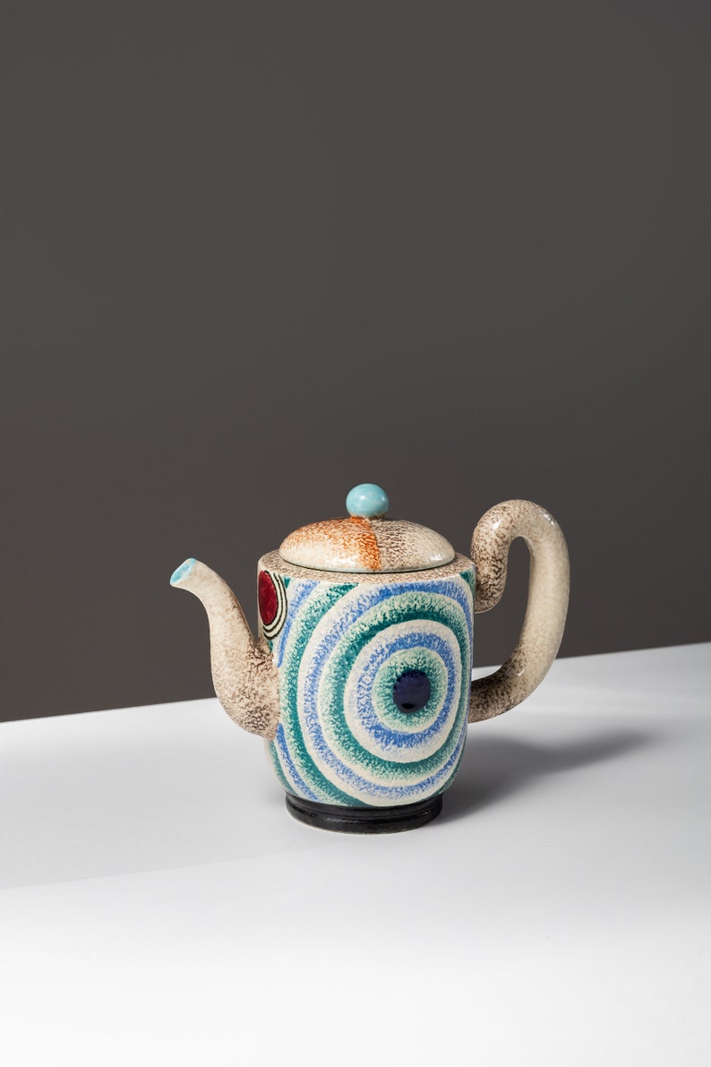 May include: A ceramic teapot with a blue, green, and white geometric pattern. The teapot has a rounded body and a curved spout. The lid has a small, round knob.