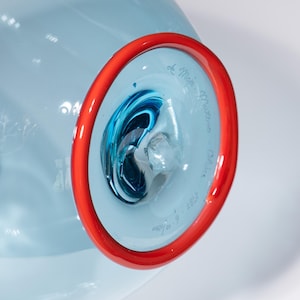 May include: A light blue glass art piece with a red rim. The interior features swirling blue and white glass. The piece is signed "de Majo Murano - Italy" and dated "1987".