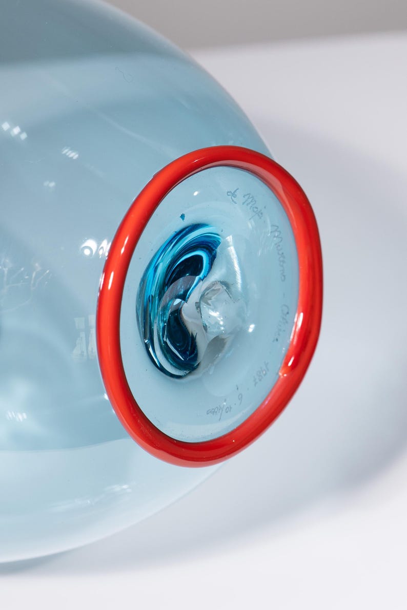 May include: A light blue glass art piece with a red rim. The interior features swirling blue and white glass. The piece is signed "de Majo Murano - Italy" and dated "1987".