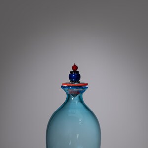 May include: A blue glass bottle with a decorative stopper. The stopper has a blue base, an orange ring, and a red and blue top. The bottle has a rounded shape and an orange ring around the base.