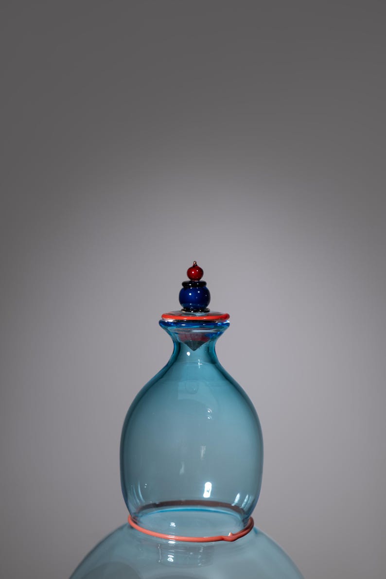 May include: A blue glass bottle with a decorative stopper. The stopper has a blue base, an orange ring, and a red and blue top. The bottle has a rounded shape and an orange ring around the base.