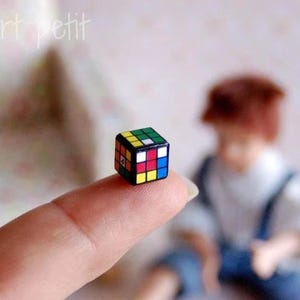 Puede incluir: Un pequeño cubo de Rubik, sostenido en un dedo. El cubo presenta la clásica disposición de cuadrados de colores: rojo, azul, verde, amarillo y blanco. El cubo mide aproximadamente 1,3 cm. El fondo está borroso, con el texto "LugArt petit" visible.