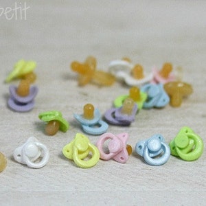 May include: A collection of miniature pacifiers in various pastel colors, including yellow, pink, blue, and purple. The pacifiers have a classic design with a round nipple and a shield. The text "LugArt petit" is in the upper left corner.