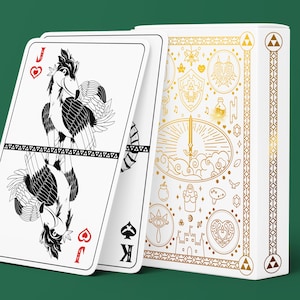 Peut inclure: Un jeu de cartes avec une boîte blanche ornée de motifs linéaires dorés représentant des symboles fantastiques, accompagné d'un valet de cœur illustré en noir et blanc d'une créature aviaire et d'un roi de pique partiellement visible.