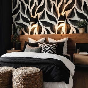 May include: A modern bedroom with a black and white leaf-patterned wallpaper headboard. The bed has white linens, black blankets, and decorative pillows. Two woven ottomans sit at the foot of the bed. Two sconces provide light.