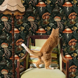 May include: An orange tabby cat stands on its hind legs, reaching towards a vintage armchair. The background features a dark wallpaper with a repeating pattern of jellyfish, fish, and mushroom-like shapes. A floor lamp with a beige shade is on the left.