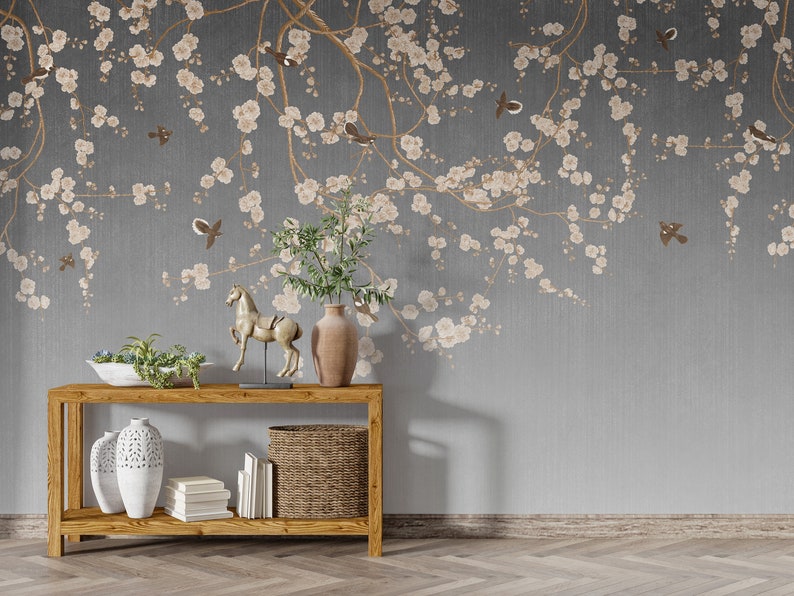May include: A wooden console table with two shelves, a white ceramic vase, a wicker basket, and a gold horse figurine. The table is in front of a gray wall with a floral wallpaper design featuring white blossoms and brown birds.