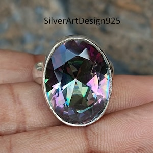 Op de afbeelding: Zilveren ring met een grote, ovaal geslepen edelsteen met een levendig kleurenspel. De facetten van de edelsteen reflecteren een spectrum aan kleuren, waaronder paars, groen en blauw. De ring is gezet in een eenvoudige, gepolijste zilveren band. De tekst "SilverArtDesign925" is zichtbaar.