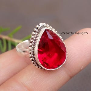 May include: A silver ring featuring a large, faceted, teardrop-shaped red gemstone. The gemstone is set in a decorative silver bezel with a beaded edge. The ring is displayed on a finger, with the text "silverArtDesign925" visible.