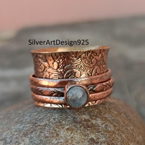 May include: A copper-colored spinner ring with a central, round, gray stone. The ring features intricate floral patterns and etched bands. The text "SilverArtDesign925" is visible on the ring.