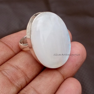 May include: A large, oval-shaped ring with a polished, white gemstone set in a silver band. The gemstone has a subtle, iridescent sheen. The ring is held against a brown background. The text "SilverArtDesign925" is visible.