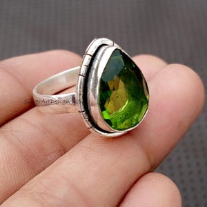 Pode incluir: Um anel de prata com uma grande pedra verde em forma de pêra. A pedra é colocada numa moldura com uma borda texturizada. O anel é segurado em uma mão, destacando a cor verde brilhante da pedra.