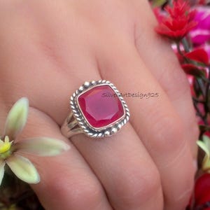 May include: A sterling silver ring featuring a large, square, ruby-red gemstone. The ring has a textured, beaded border around the stone and a split shank. The ring is on a hand, with flowers in the background. The text "SilverArtDesign925" is visible.