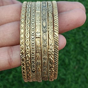 May include: A set of seven gold-toned bangles with intricate engraved designs. The bangles are stacked on top of each other and have a variety of patterns, including floral, geometric, and abstract.