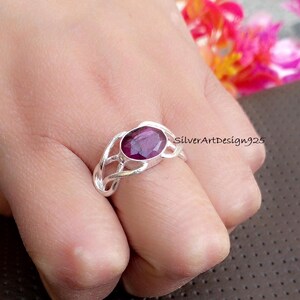May include: A silver ring featuring an oval, deep purple gemstone. The ring has a woven design around the gemstone and is worn on a finger. The text "SilverArtDesign925" is visible on the right side of the image.
