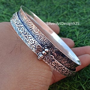 May include: A silver bangle bracelet featuring a floral design and a bee charm. The bracelet is made of two bands that spin around each other.