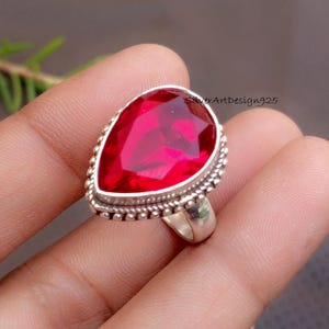 May include: A silver ring featuring a large, pear-shaped, vibrant red gemstone. The gemstone is set in a silver band with a beaded border. The ring is a statement piece, perfect for adding a pop of color.