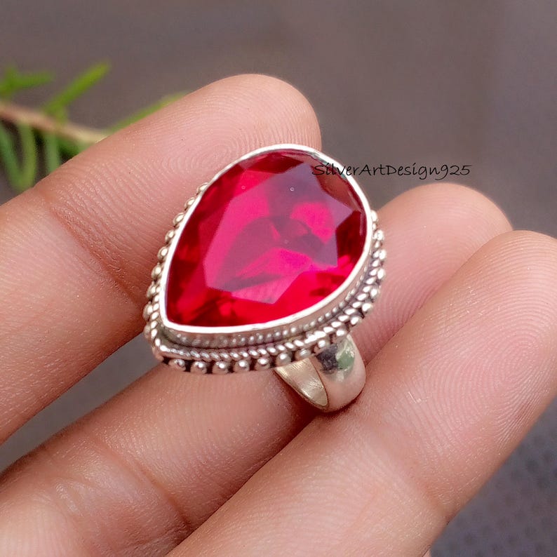 May include: A silver ring featuring a large, pear-shaped, vibrant red gemstone. The gemstone is set in a silver band with a beaded border. The ring is a statement piece, perfect for adding a pop of color.